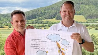 Zwei Männer lächeln und halten ein Blatt Papier mit deutschem Text, Zeichnungen und einem Bleistift. Einer trägt ein rotes Shirt, der andere ein weißes Polo. Hinter ihnen ist eine Berglandschaft.