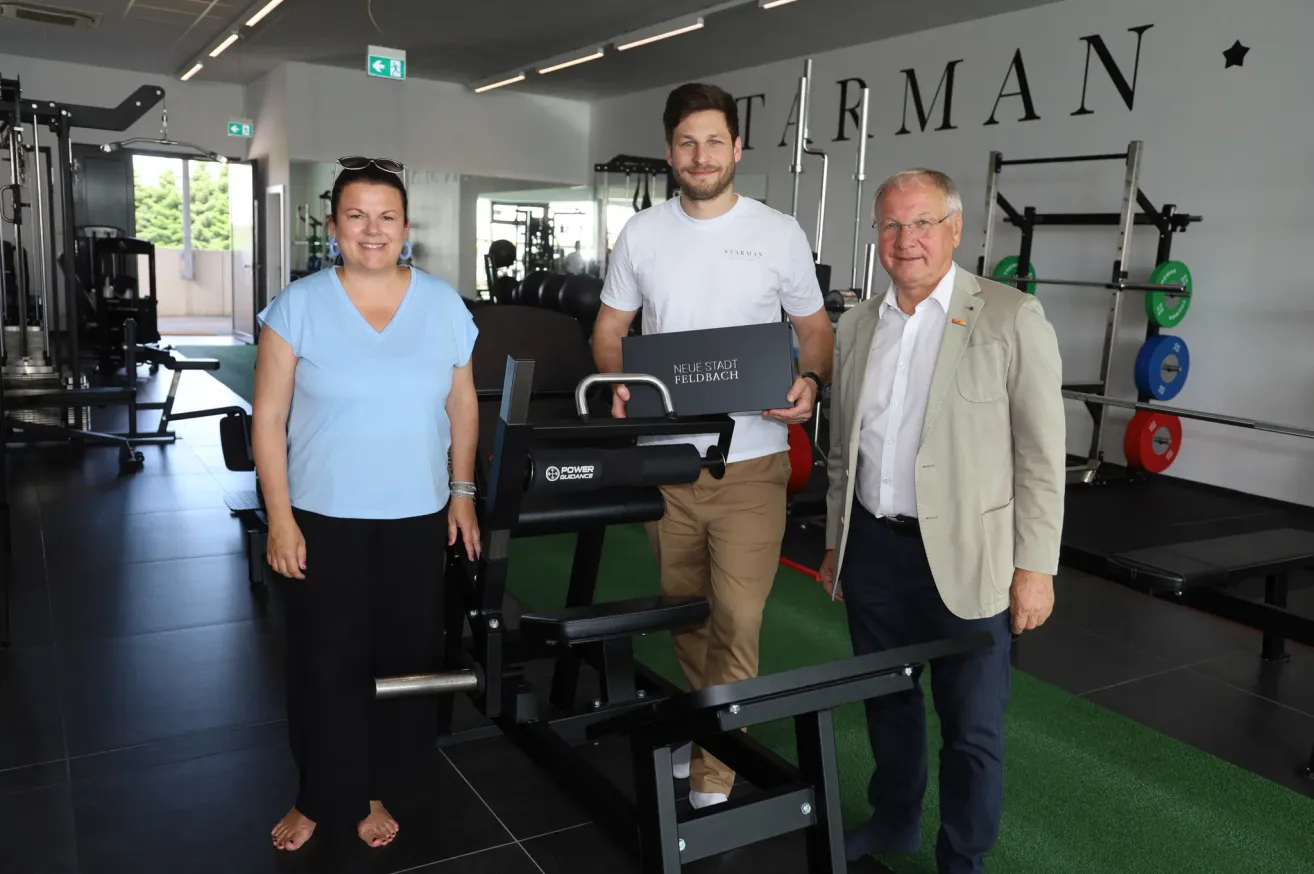 Drei Personen stehen in einem Fitnessstudio und halten eine schwarze Box. Der Mann in der Mitte lächelt, während der Mann rechts eine Brille trägt. Hinter ihnen ist Fitnessequipment zu sehen.
