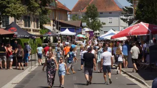 Eine belebte Straße mit einer Menschenmenge, die an einem sonnigen Tag läuft. Es gibt Geschäfte, Zelte und Bäume auf beiden Seiten. Die Menschen gehen in verschiedene Richtungen.