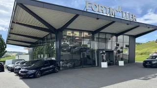 Das Fortmüller Autohaus zeigt eine moderne Glasfassade mit geparkten Autos davor. Das Gebäude befindet sich unter einem großen Vordach, das den Namen des Autohauses zeigt.