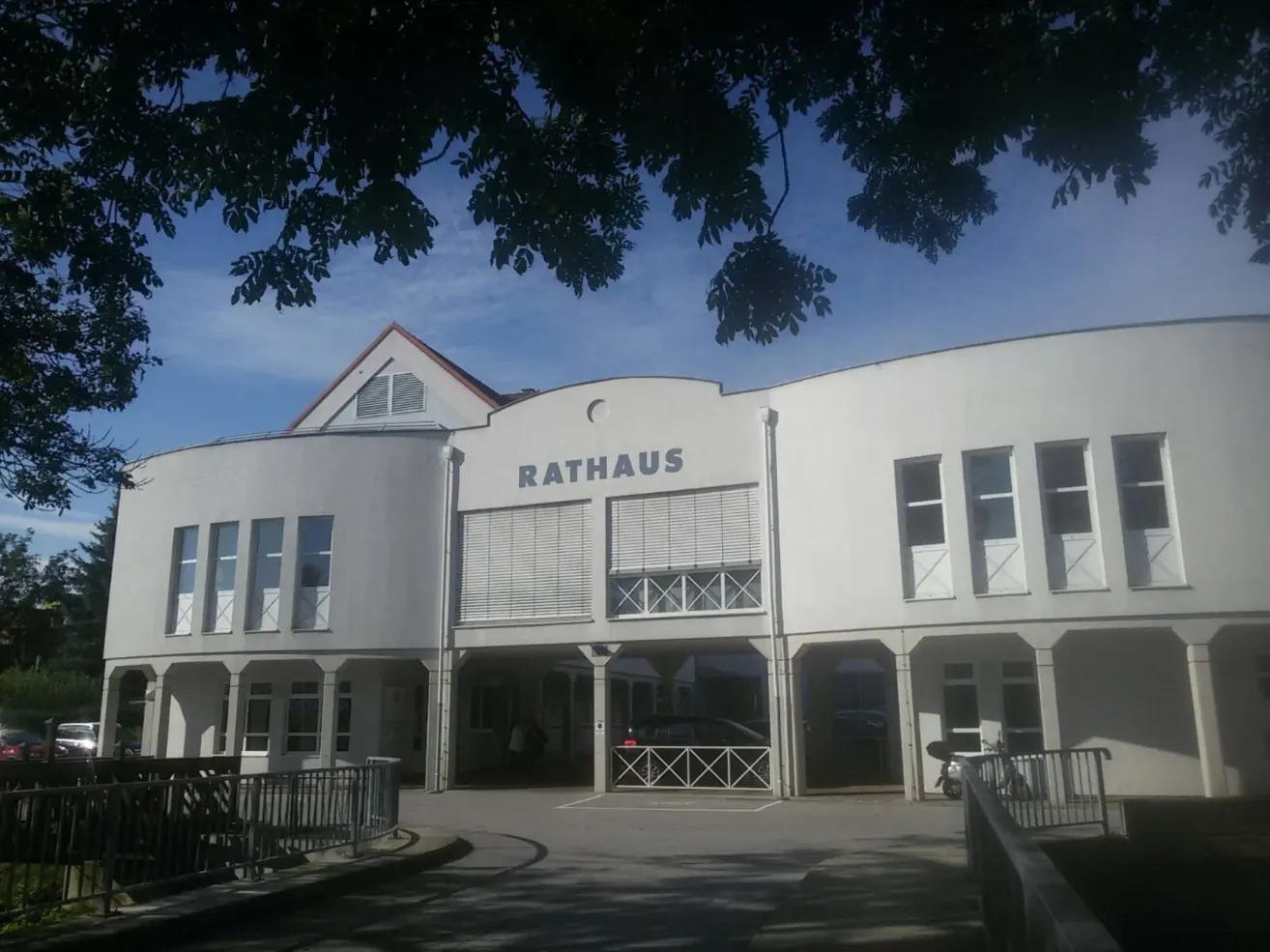 Ein weißes Gebäude mit der Aufschrift Rathaus ist zu sehen. Es hat viele Fenster und ist von Metallgeländern umgeben. Ein Auto ist vorn geparkt, und es gibt einen Baum in der Nähe.