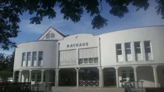 Ein weißes Gebäude mit der Aufschrift Rathaus ist zu sehen. Es hat viele Fenster und ist von Metallgeländern umgeben. Ein Auto ist vorn geparkt, und es gibt einen Baum in der Nähe.