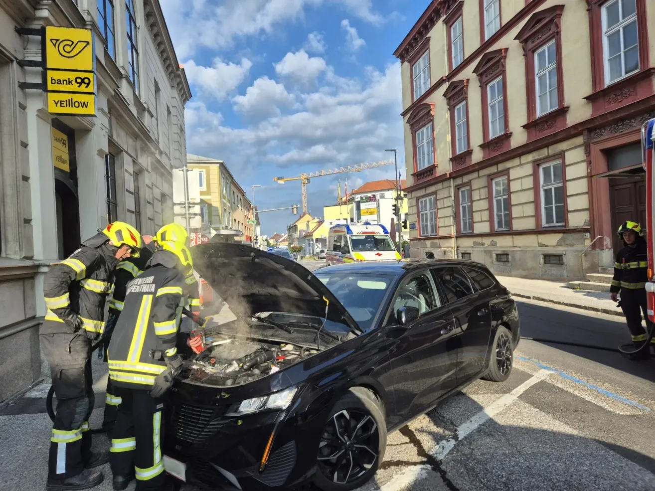 Drei Feuerwehrleute inspizieren ein schwarzes Auto mit offenem Motorhaube. In der Nähe steht ein weißer Van mit einem roten Kreuz auf der Seite.