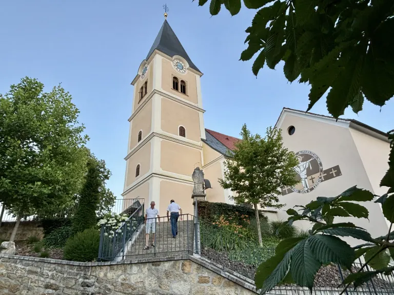 Zwei Männer steigen die Stufen zu einer Kirche hinauf, deren Turm mit einer Uhr geschmückt ist. Das Gebäude ist von üppigem Grün und Bäumen umgeben.