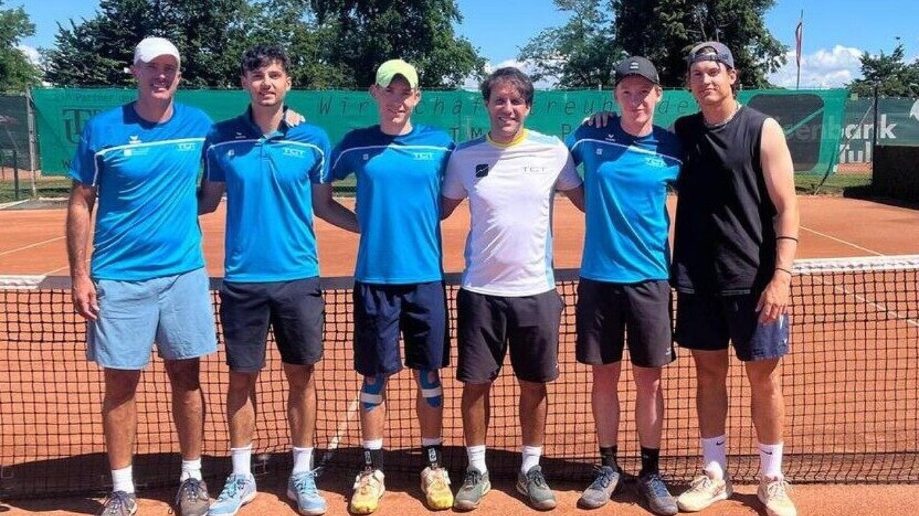 Fünf Männer in blauen und weißen Trikots stehen lächelnd auf einem Tennisplatz. Sie tragen Turnschuhe und Kniestützen. Ein Zaun und eine grüne Wand mit Text stehen hinter ihnen.