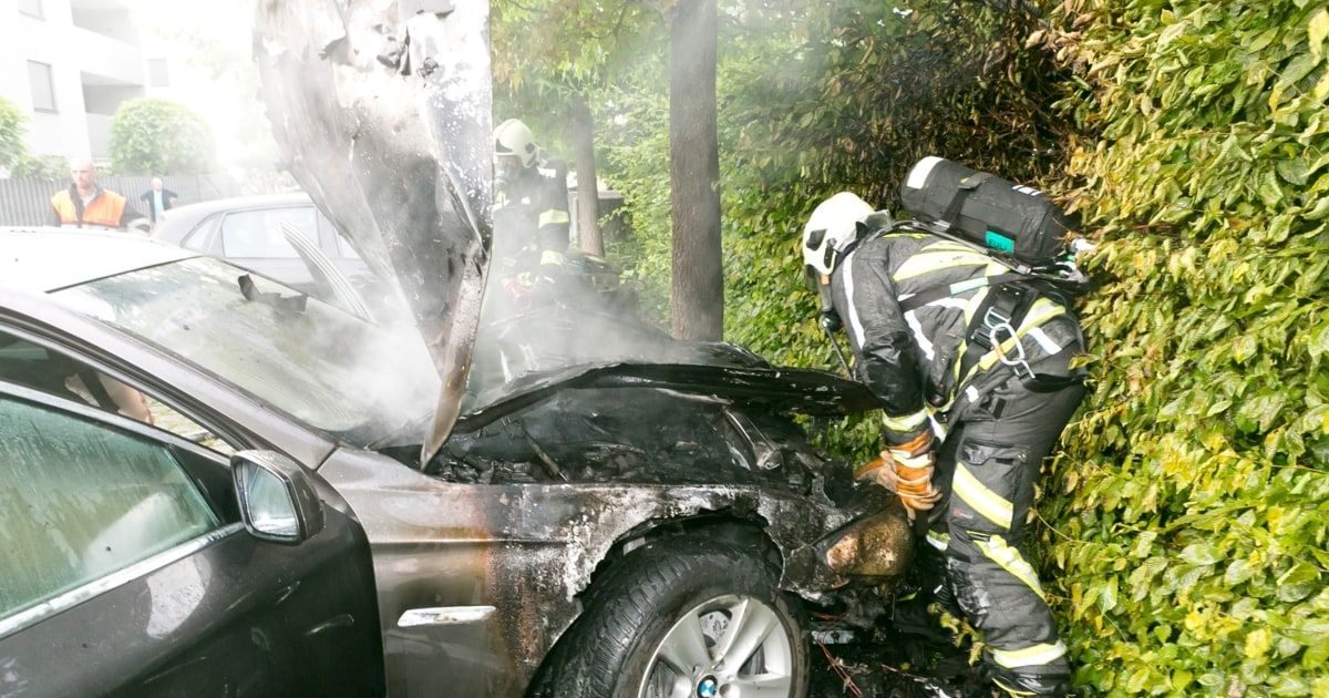 Feuerwehrleute untersuchen ein verbranntes Auto, wobei ein Feuerwehrmann das Fahrzeug inspiziert und ein anderer in der Nähe steht.