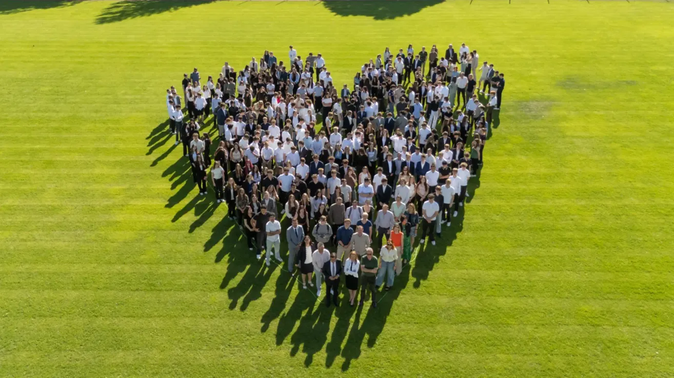 Eine große Gruppe von Menschen steht in Herzform auf einem grünen Feld unter klarem Himmel. Einige tragen formelle Kleidung, andere lässige Kleidung.