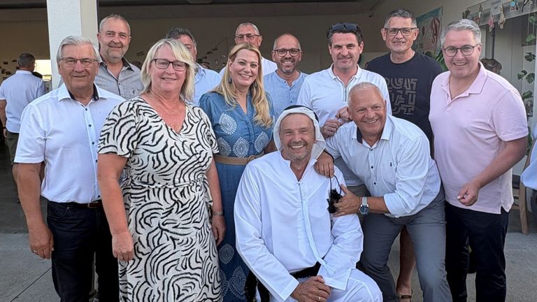 Eine Gruppe von Erwachsenen in formeller Kleidung lächelt und posiert für ein Foto. Der Mann in der Mitte sitzt und hält eine Kamera.