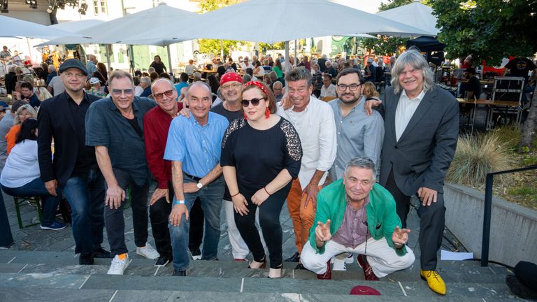 Eine Gruppe von Männern und einer Frau steht gemeinsam auf Stufen und lächelt für ein Foto. Dahinter sitzen und stehen Menschen in einem überfüllten Außenbereich.