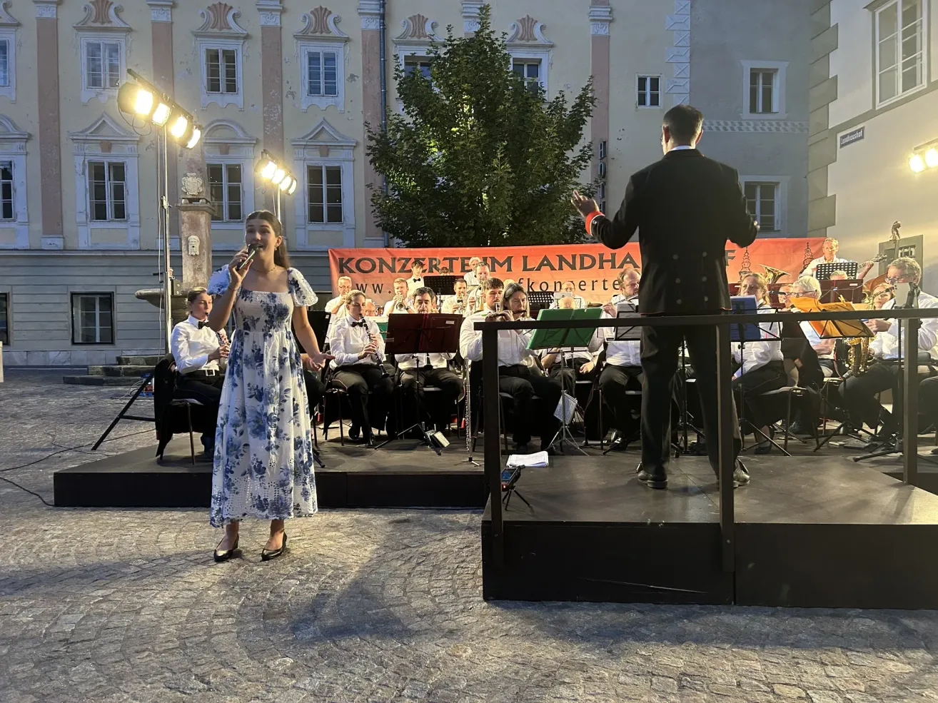 Eine Frau in einem Blumenkleid singt in ein Mikrofon auf der Bühne, während eine Band hinter ihr spielt. Die Bühne ist mit einem Banner für Konzerte im Landhaus geschmückt.