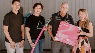 Zwei Erwachsene, ein Mann und eine Frau, stehen nebeneinander vor einer Wand. Der Mann hält eine rosa Box und die Frau eine rosa Röhre. Sie lächeln und posieren für ein Foto.