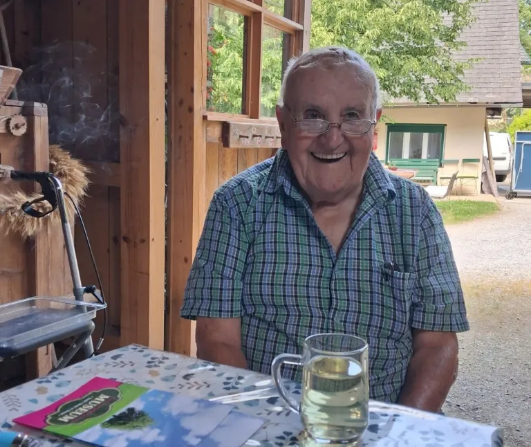 Ein älterer Mann mit Brille sitzt lächelnd vor einem Holzhaus. Er sitzt an einem Tisch mit einem Glas Getränk und einem Buch. Hinter ihm befindet sich eine Rasenfläche mit Bäumen und einem Gebäude.