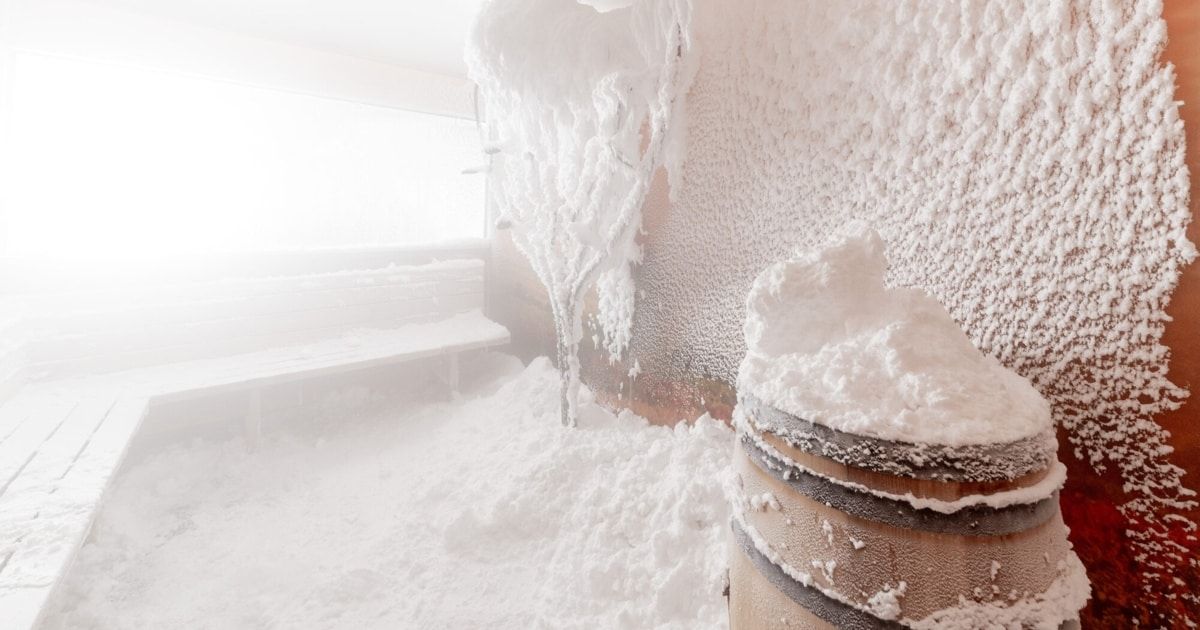 Ein schneebedeckter Raum mit einem hölzernen Fass und einer Bank, die mit Schnee bedeckt sind. Die Wände und die Decke sind ebenfalls mit Schnee bedeckt.