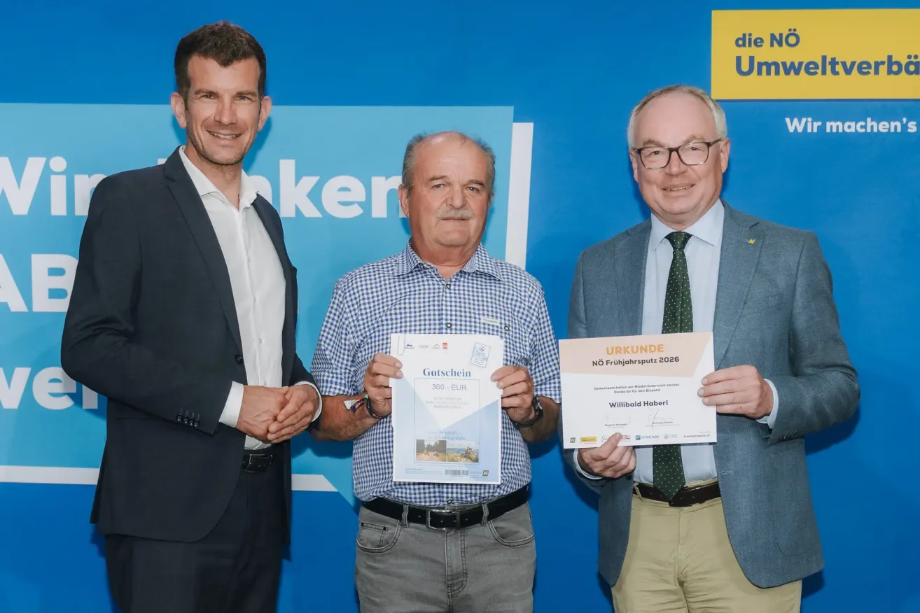 Drei Männer stehen nebeneinander vor einem blauen Hintergrund. Der Mann in der Mitte hält ein Zertifikat mit der Aufschrift 'Gutschein' und '300 EUR.' Der Mann auf der rechten Seite hält ein Zertifikat mit der Aufschrift 'Urkunde' und 'NÖ Freijahrssputz 2026.' Sie tragen alle formale Kleidung.