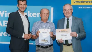 Drei Männer stehen nebeneinander vor einem blauen Hintergrund. Der Mann in der Mitte hält ein Zertifikat mit der Aufschrift 'Gutschein' und '300 EUR.' Der Mann auf der rechten Seite hält ein Zertifikat mit der Aufschrift 'Urkunde' und 'NÖ Freijahrssputz 2026.' Sie tragen alle formale Kleidung.
