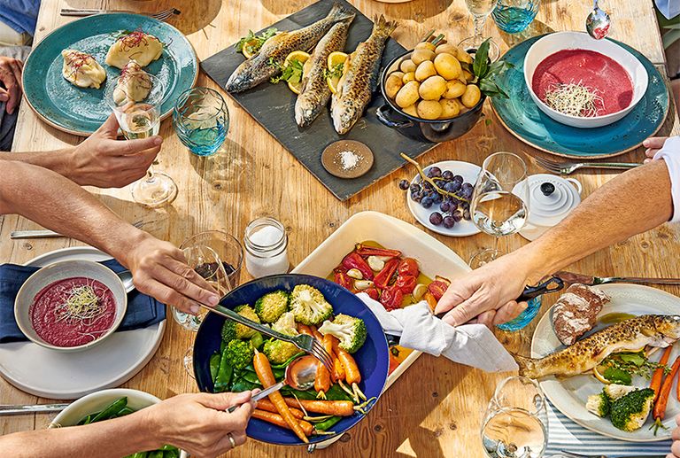 Ein Holztisch mit verschiedenen Tellern, Schüsseln und Gerichten, darunter Fisch, Gemüse und eine rote Suppe. Mehrere Hände greifen nach den Gerichten.