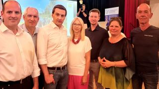 Fünf Personen stehen eng für ein Foto in einem Konferenzraum. Eine Frau in der Mitte trägt eine Brille und eine Halskette. Der Mann auf der linken Seite hat einen Gürtel. Ein Mann im Hintergrund hält eine Kamera.