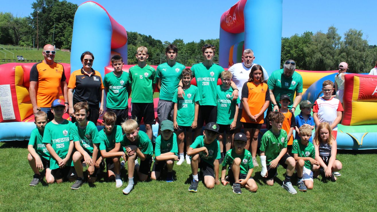 Eine Jugendsportmannschaft in grünen Trikots, die vor einem aufblasbaren Schloss lächelnd und posierend steht, mit Erwachsenen hinter ihnen. Sie sind im Freien auf einem Rasenfeld mit Bäumen im Hintergrund.