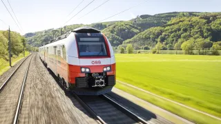 Ein weißer und roter Zug mit den Buchstaben OBB auf der Vorderseite rast auf einer Bahnstrecke, umgeben von grünen Feldern.