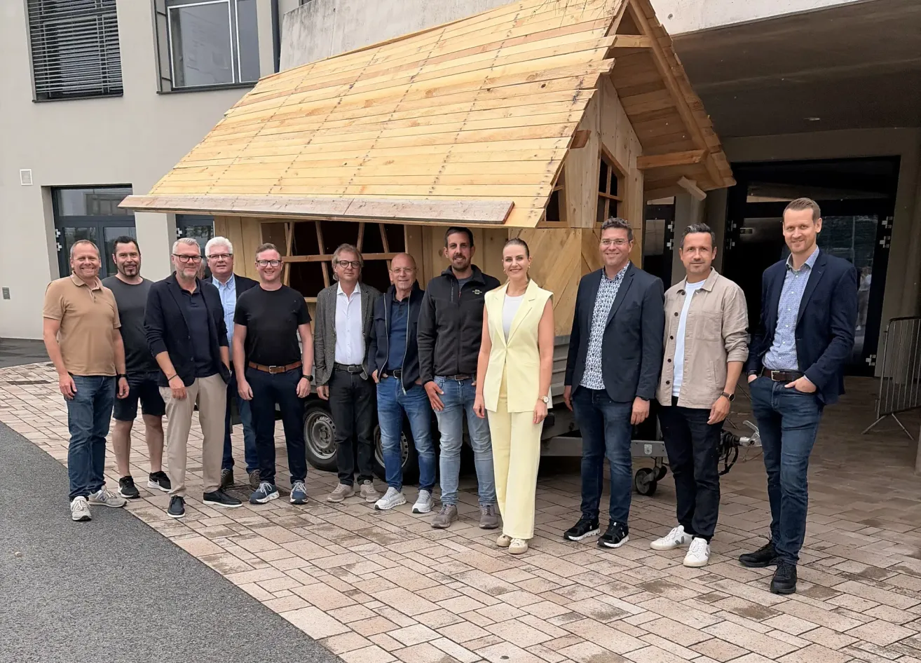 Eine Gruppe von Männern und einer Frau steht vor einem kleinen Holzhaus. Sie tragen formelle Kleidung. Das Haus hat ein schräges Dach und ist an einen Anhänger angeschlossen.