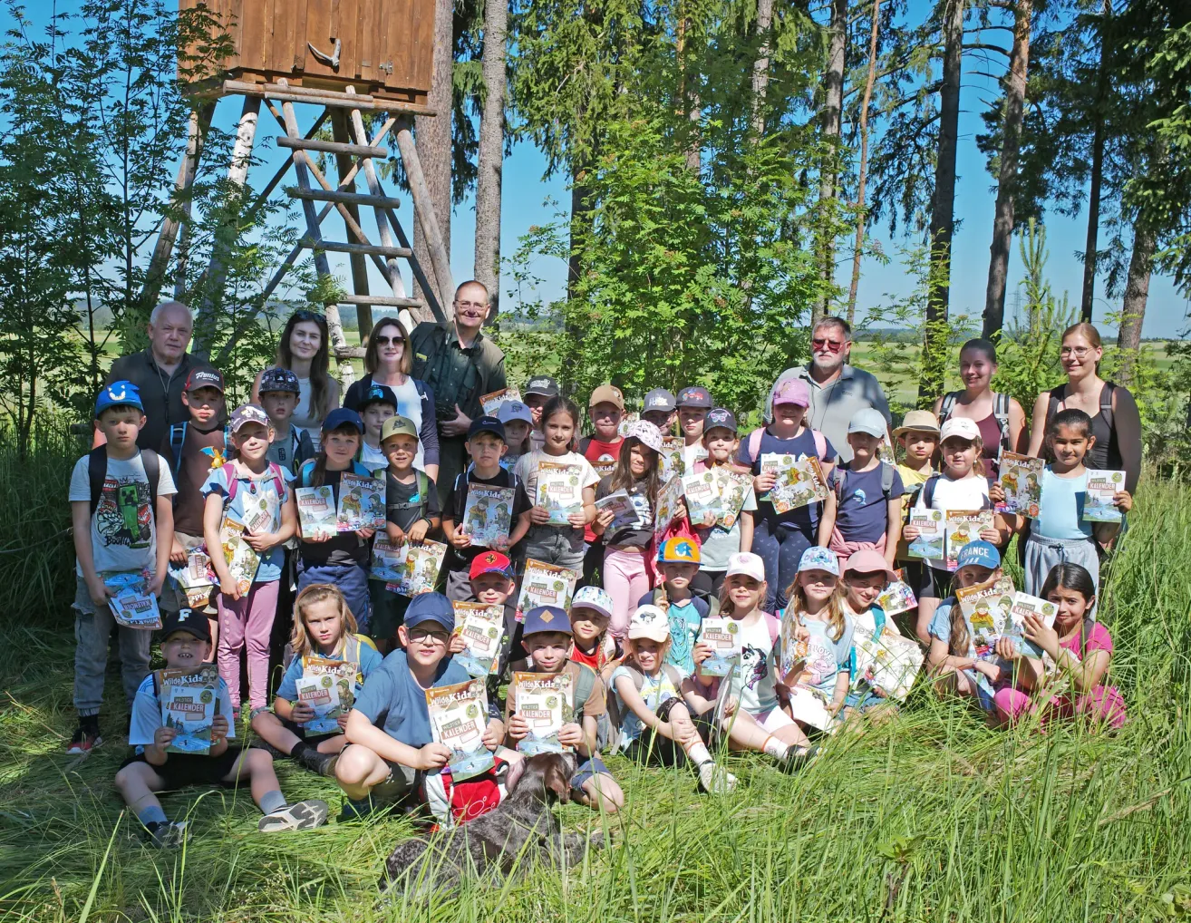 Eine Gruppe von Kindern und Erwachsenen posiert für ein Foto im Wald mit einem Holzturm im Hintergrund, alle halten Urkunden und tragen Hüte und Brillen.