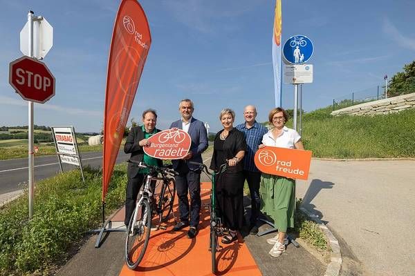 Eine Gruppe von Menschen steht auf einem Fahrradweg, hält ein Schild mit der Aufschrift 'Radfreude' hoch und posiert für ein Foto. Sie stehen neben einem Stoppschild und einer Fahne.