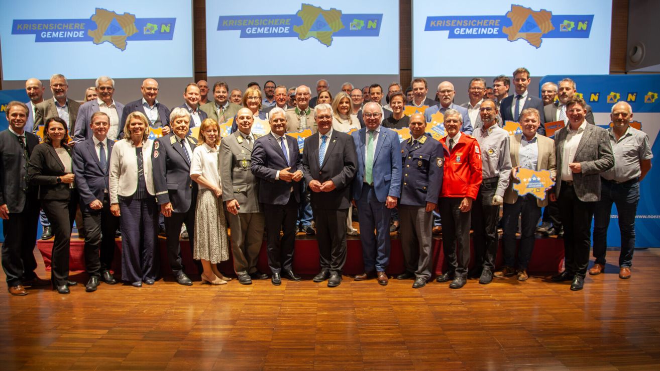 Eine Gruppe von Menschen in formeller Kleidung, einschließlich Anzüge und Krawatten, steht auf einer Bühne, wahrscheinlich für eine Veranstaltung oder Preisverleihung. Dahinter befinden sich Bildschirme mit Logos und Text.