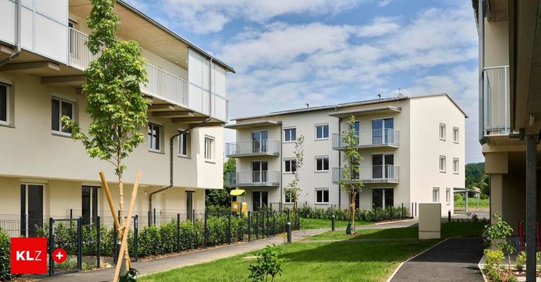 Eine Reihe moderner weißer Mehrfamilienhäuser mit Balkonen, einem kleinen Park mit Spielplatz und einem Gehweg, umgeben von Büschen und Bäumen unter einem bewölkten Himmel.
