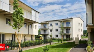 Eine Reihe moderner weißer Mehrfamilienhäuser mit Balkonen, einem kleinen Park mit Spielplatz und einem Gehweg, umgeben von Büschen und Bäumen unter einem bewölkten Himmel.
