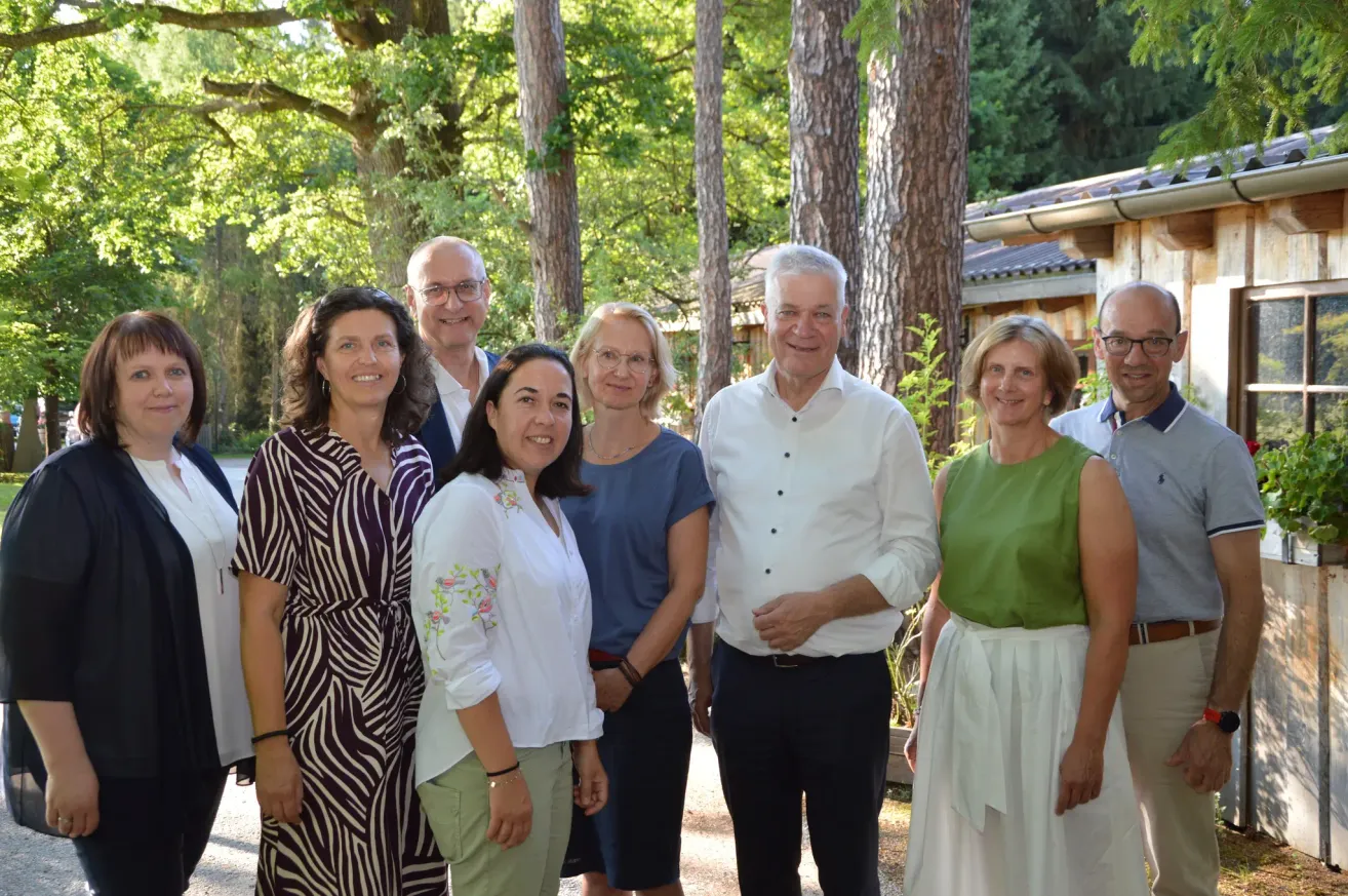 Eine Gruppe Erwachsener, lässig gekleidet, steht gemeinsam in einem Garten mit Bäumen und einem Gebäude im Hintergrund und posiert für ein Foto.