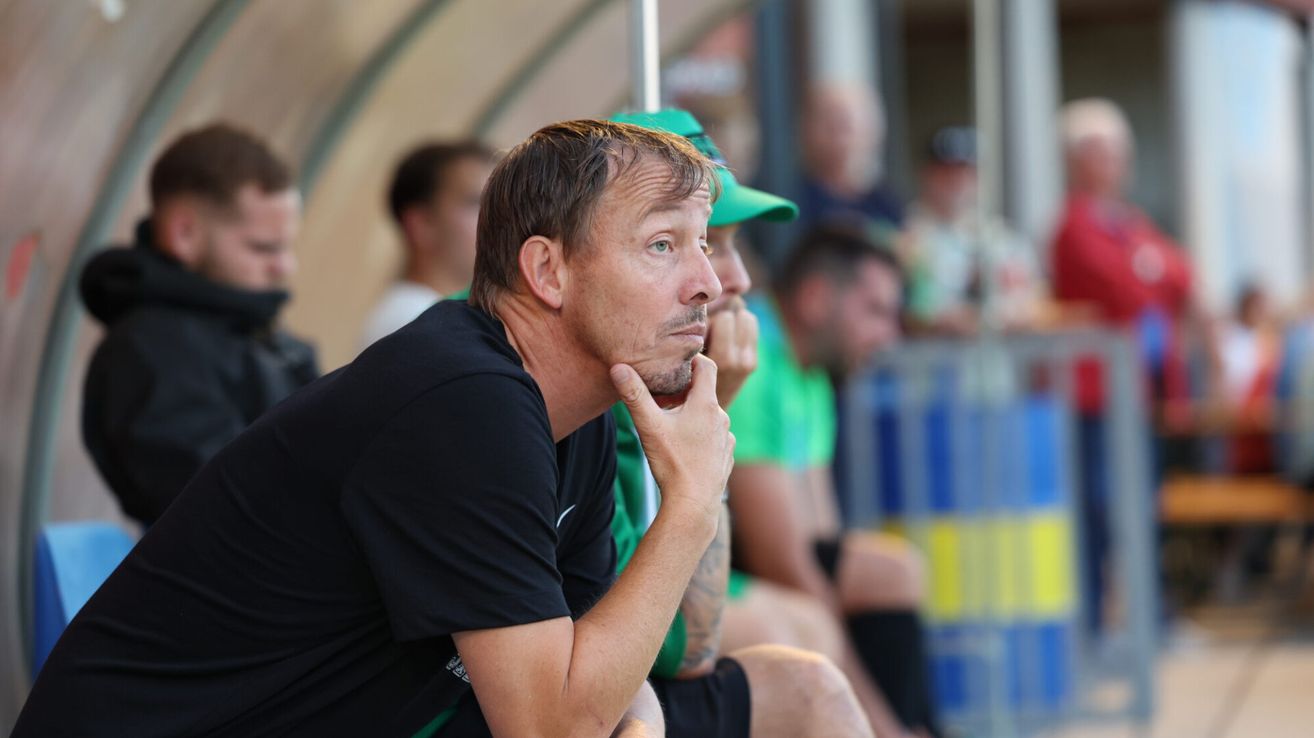 Ein Mann mit schwarzem T-Shirt sitzt mit der Hand am Kinn und beobachtet ein Fußballspiel. Hinter ihm sieht ein Mann in einem grünen Shirt und einer grünen Kappe das Spiel.