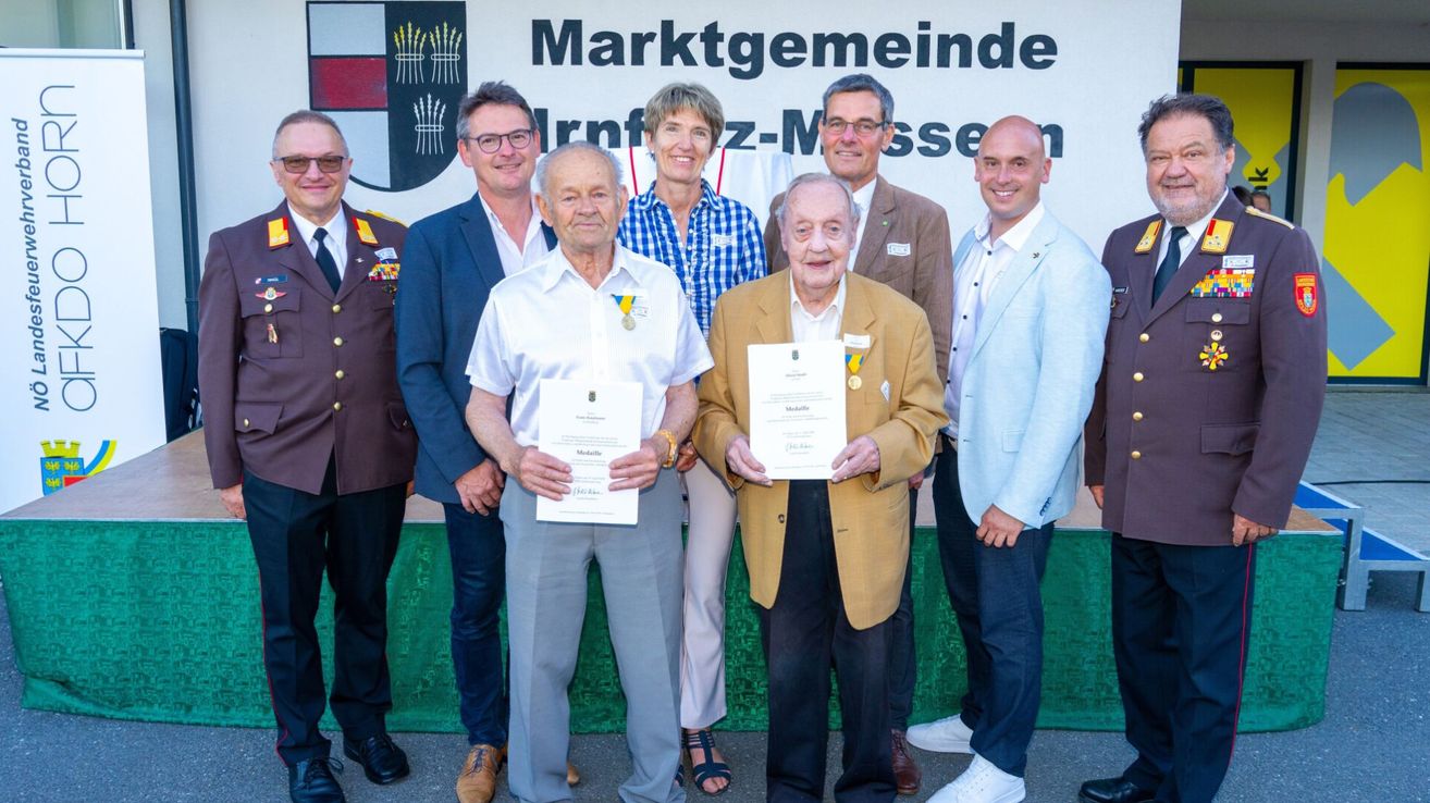 Zwei ältere Männer halten Urkunden vor einem Schild, auf dem 'Marktgemeinde Irnfritz-Messen' steht. Sie sind von anderen Männern und Frauen umgeben, alle feierlich gekleidet. Sie scheinen bei einer Preisverleihung zu sein.