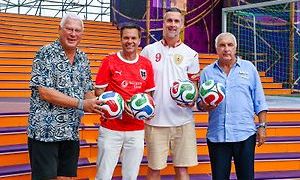 Vier Männer stehen auf einer Bühne mit bunten Stufen und halten Fußballbälle. Ein Mann trägt ein rotes Trikot mit Logo, ein anderer trägt ein weißes Trikot mit der Nummer 9. Im Hintergrund befindet sich eine farbenfrohe Bühnendekoration und ein großer Torpfosten.