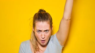 Eine Frau mit blonden Haaren in einem grauen T-Shirt sieht schockiert aus, als sie gegen eine gelbe Wand einen Tablet-Computer ansieht.