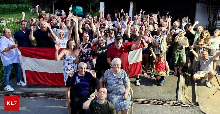 Eine Gruppe von Menschen, einschließlich älterer Frauen, posiert für ein Foto, während sie Flaggen hochhalten und jubeln. Ein Mann hält im Vordergrund eine Flagge.