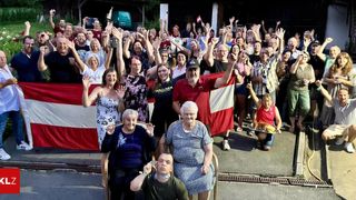 Eine Gruppe von Menschen, einschließlich älterer Frauen, posiert für ein Foto, während sie Flaggen hochhalten und jubeln. Ein Mann hält im Vordergrund eine Flagge.