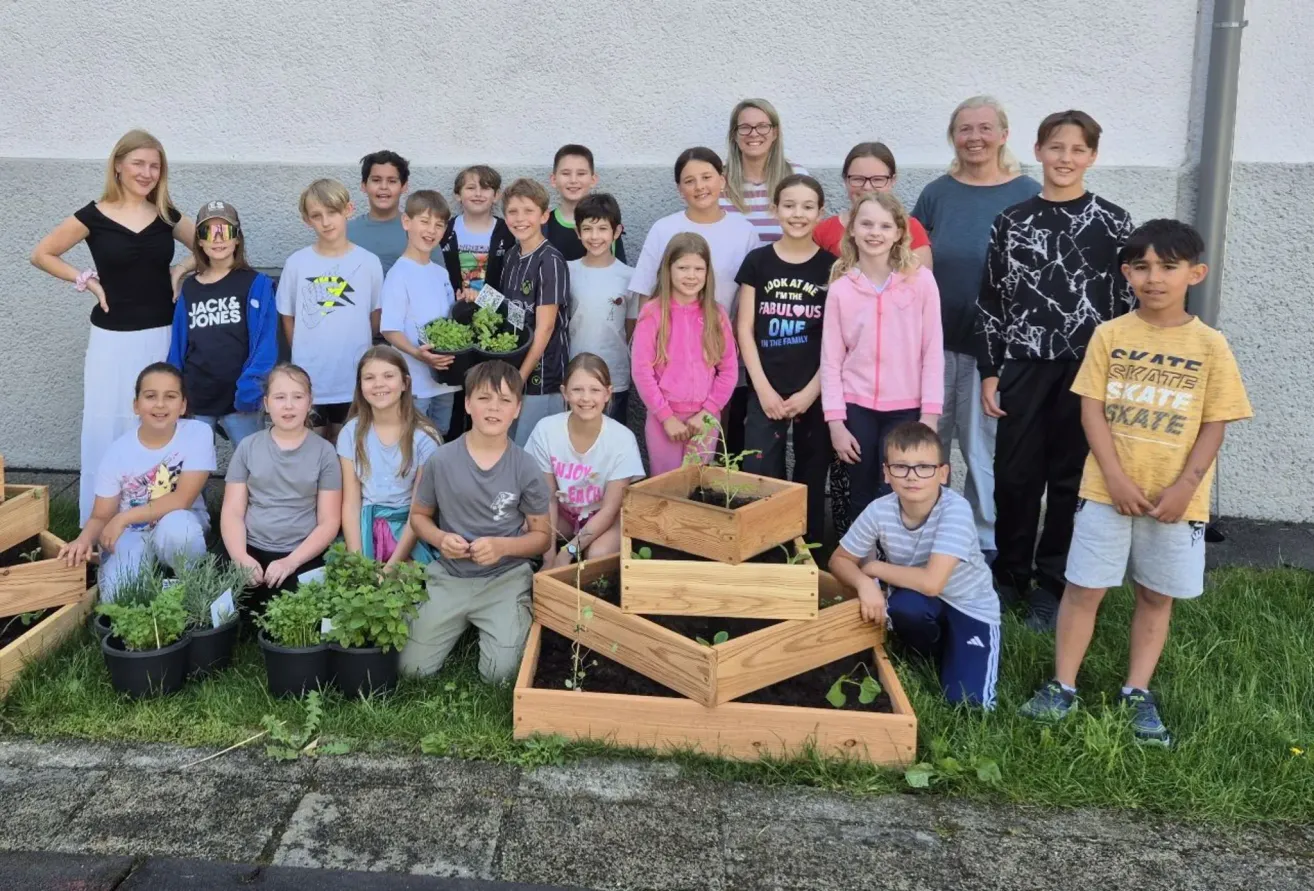Eine Gruppe von Kindern und Erwachsenen posiert für ein Foto im Freien, hält Pflanzen und Töpfe. Im Vordergrund steht ein dreistufiger hölzerner Pflanzkasten gefüllt mit Grün. Die Kinder lächeln und scheinen die Aktivität zu genießen.