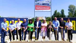 Eine Gruppe von Menschen steht vor einem Gebäudeschild für Reihenhauser. Sie halten Schaufeln für eine Grundsteinlegungszeremonie.