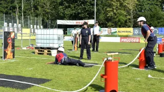 Auf einem Feld liegt eine Person mit weißem Helm auf dem Boden, während andere stehen, darunter eine mit Mütze und Uhr. Ein roter Feuerlöscher ist in der Nähe.
