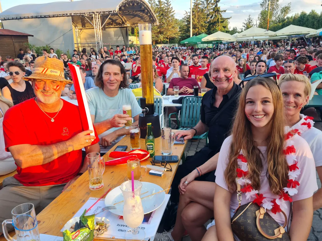 Drei Männer und eine Frau sitzen an einem Tisch mit Getränken und Snacks. Sie lächeln und posieren für ein Foto mit einem großen Becher in der Mitte. Im Hintergrund ist eine Menschenmenge unter Schirmen sitzend zu sehen.