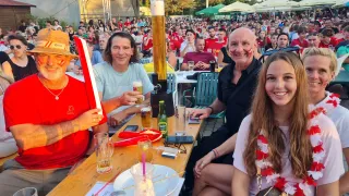 Drei Männer und eine Frau sitzen an einem Tisch mit Getränken und Snacks. Sie lächeln und posieren für ein Foto mit einem großen Becher in der Mitte. Im Hintergrund ist eine Menschenmenge unter Schirmen sitzend zu sehen.