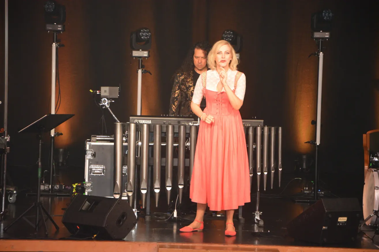 Eine Frau in einem roten Kleid steht auf der Bühne, wahrscheinlich bei einer Aufführung. Hinter ihr spielt ein Mann ein Yamaha-Klavier, mit einem Scheinwerfer auf ihn gerichtet. Die Bühne ist mit Lautsprechern, einem Koffer und einem großen Musikinstrument ausgestattet.
