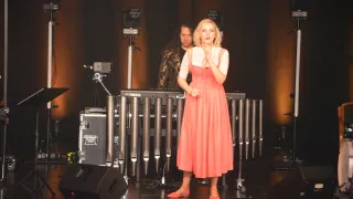 Eine Frau in einem roten Kleid steht auf der Bühne, wahrscheinlich bei einer Aufführung. Hinter ihr spielt ein Mann ein Yamaha-Klavier, mit einem Scheinwerfer auf ihn gerichtet. Die Bühne ist mit Lautsprechern, einem Koffer und einem großen Musikinstrument ausgestattet.