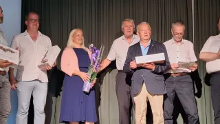 Eine Frau mit blonden Haaren, die einen Blumenstrauß hält, steht neben zwei älteren Männern vor einem Vorhang. Der Mann auf der rechten Seite hält eine weiße Schachtel.