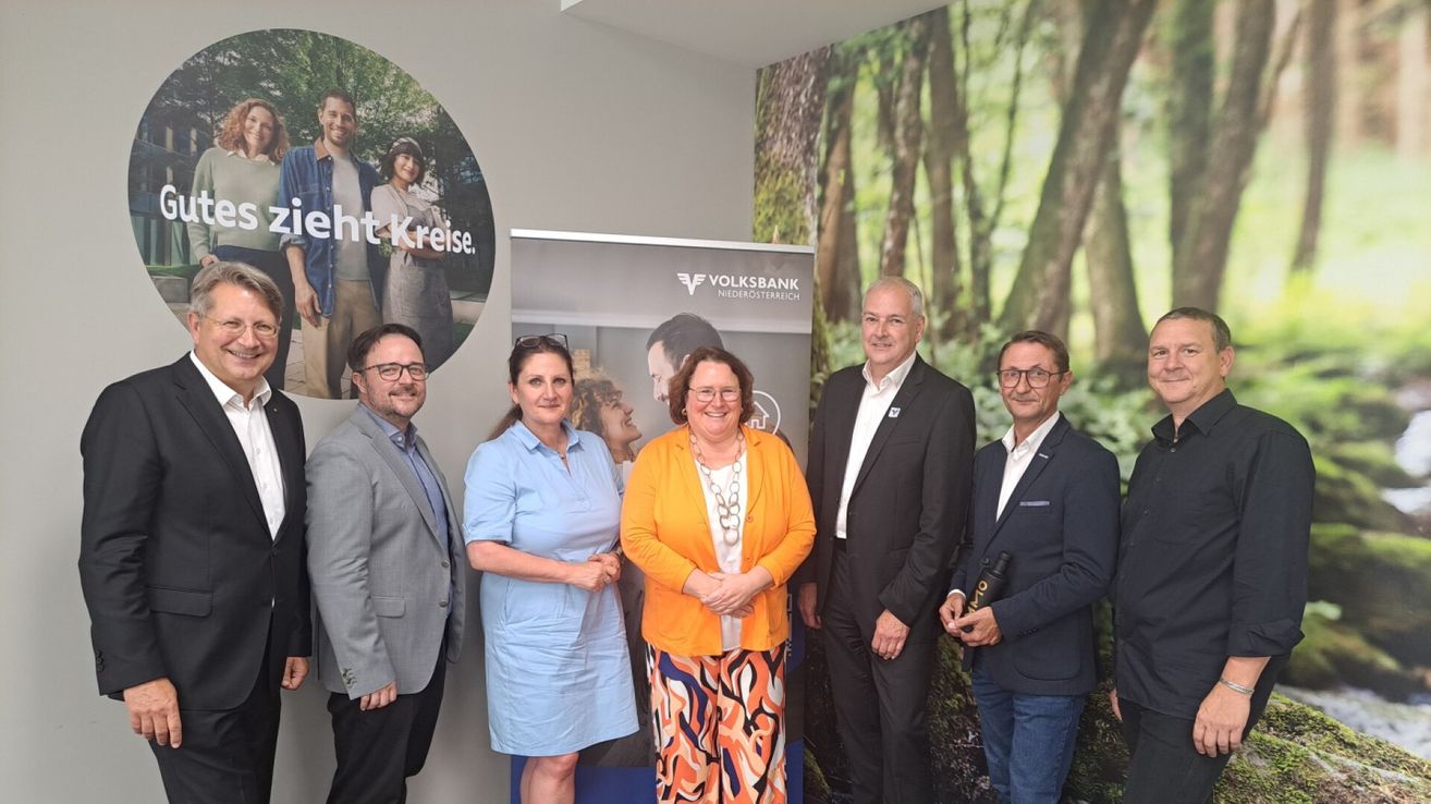 Fünf Menschen stehen lächelnd zusammen in einem Raum. Einer hält eine Flasche. Ein Banner hinter ihnen lautet 'Volksbank Niederosterreich'. Ein kreisförmiges Bild an der Wand zeigt eine Familie.