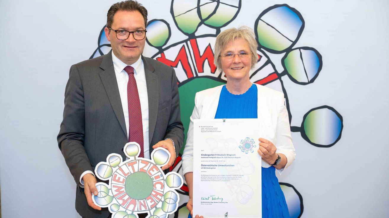 Ein Mann und eine Frau stehen zusammen und halten eine Urkunde. Der Mann trägt einen Anzug und eine Krawatte, und die Frau trägt ein blaues Kleid. Hinter ihnen ist eine bunte Wand mit einem Logo.