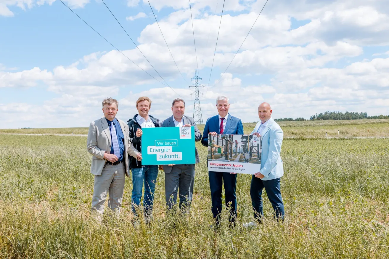 Fünf Männer in Anzügen stehen in einem Grasfeld und halten Schilder. Der Mann in der Mitte hält ein grünes Schild mit der Aufschrift 'Wir bauen Energie-zukunft'. Hinter ihm hält ein anderer Mann ein weißes Schild mit einem Gebäude darauf. Hinter den Schildern reichen Stromleitungen bis in die Ferne.