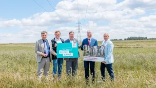Fünf Männer in Anzügen stehen in einem Grasfeld und halten Schilder. Der Mann in der Mitte hält ein grünes Schild mit der Aufschrift 'Wir bauen Energie-zukunft'. Hinter ihm hält ein anderer Mann ein weißes Schild mit einem Gebäude darauf. Hinter den Schildern reichen Stromleitungen bis in die Ferne.