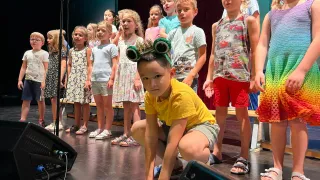 Kinder führen auf einer Bühne auf; ein Junge vorne trägt eine Krone und schaut in die Kamera. Dahinter steht eine Gruppe von Kindern in einer Reihe, einige tragen Sandalen. Ein Mikrofon steht links auf einem Ständer.