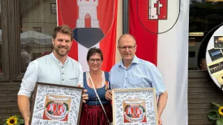 Drei Menschen stehen draußen und halten signierte gerahmte Urkunden, lächelnd. Hinter ihnen befindet sich ein Gebäude mit einer Flagge, die eine Burg und ein Kreuz zeigt.
